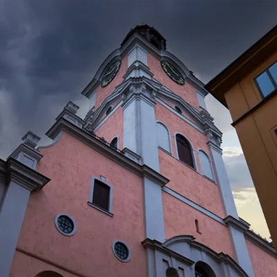 Rosa kyrktorn mot mörk dramatisk himmel och intilliggande hus.